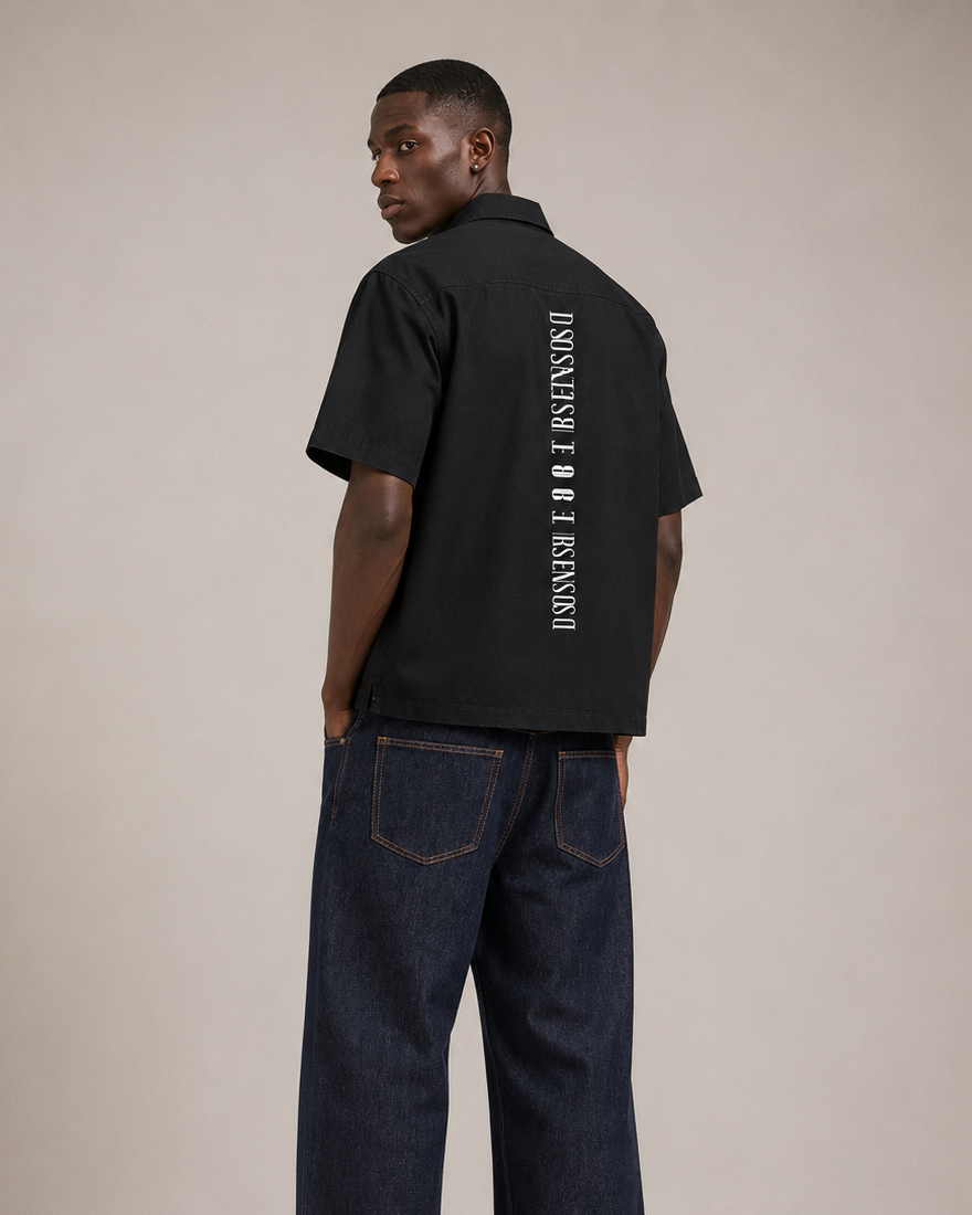 Man wearing a black shirt and dark jeans with brand logos on a beige background