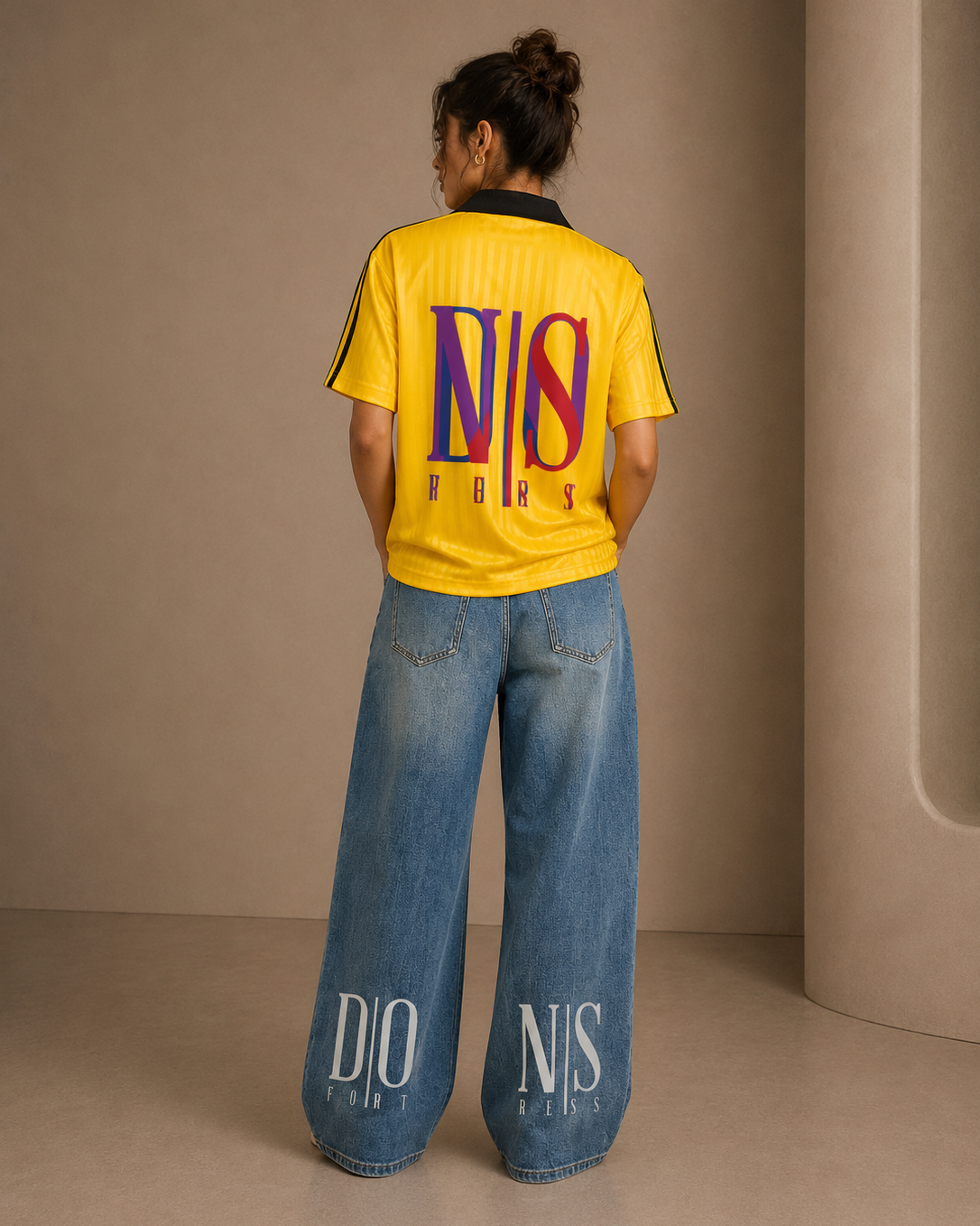 Person wearing a yellow t-shirt with colorful branding and blue jeans with text, standing against a beige wall.