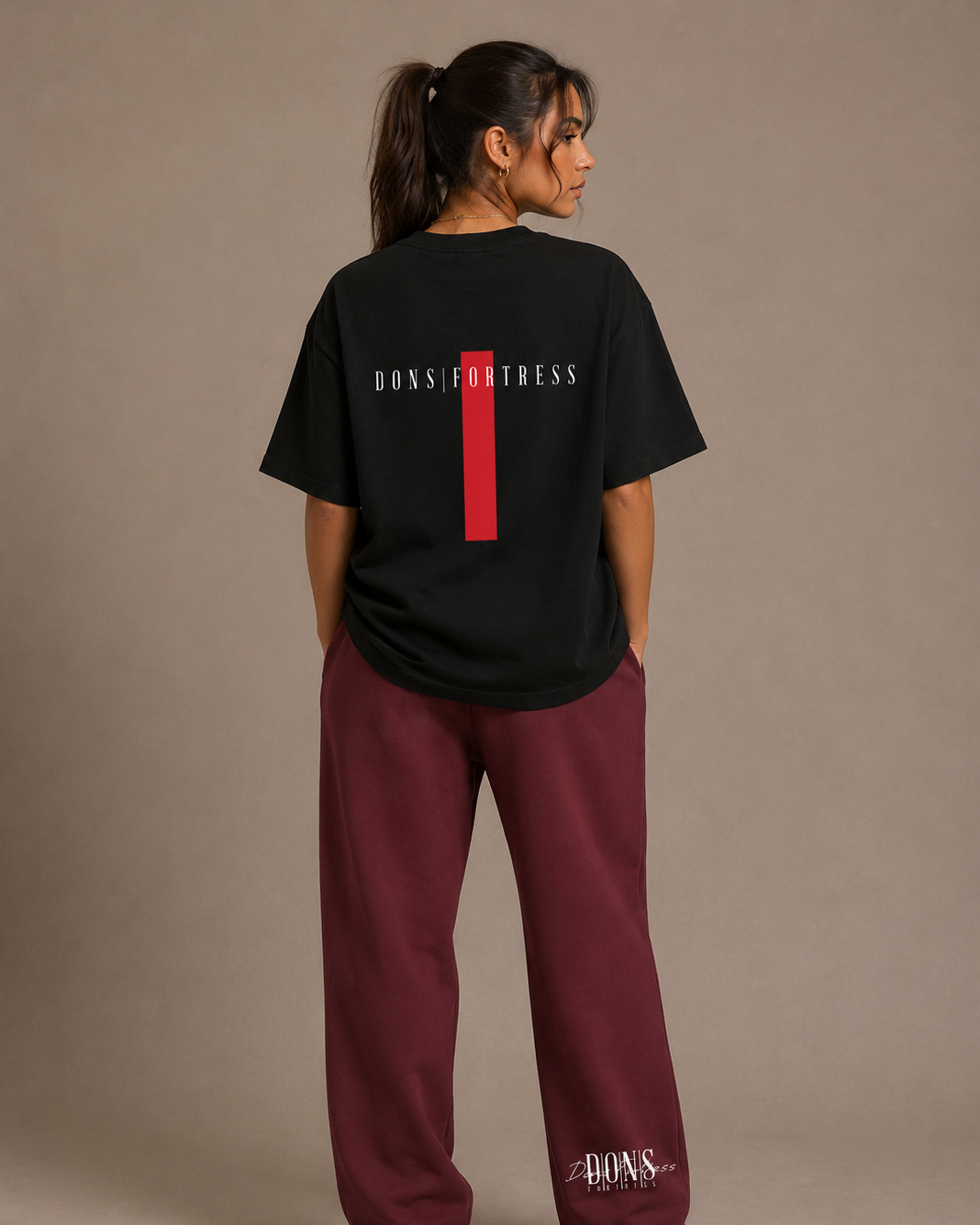 Person wearing a black t-shirt with branding on the back and maroon pants with branding on the side, standing against a beige background.