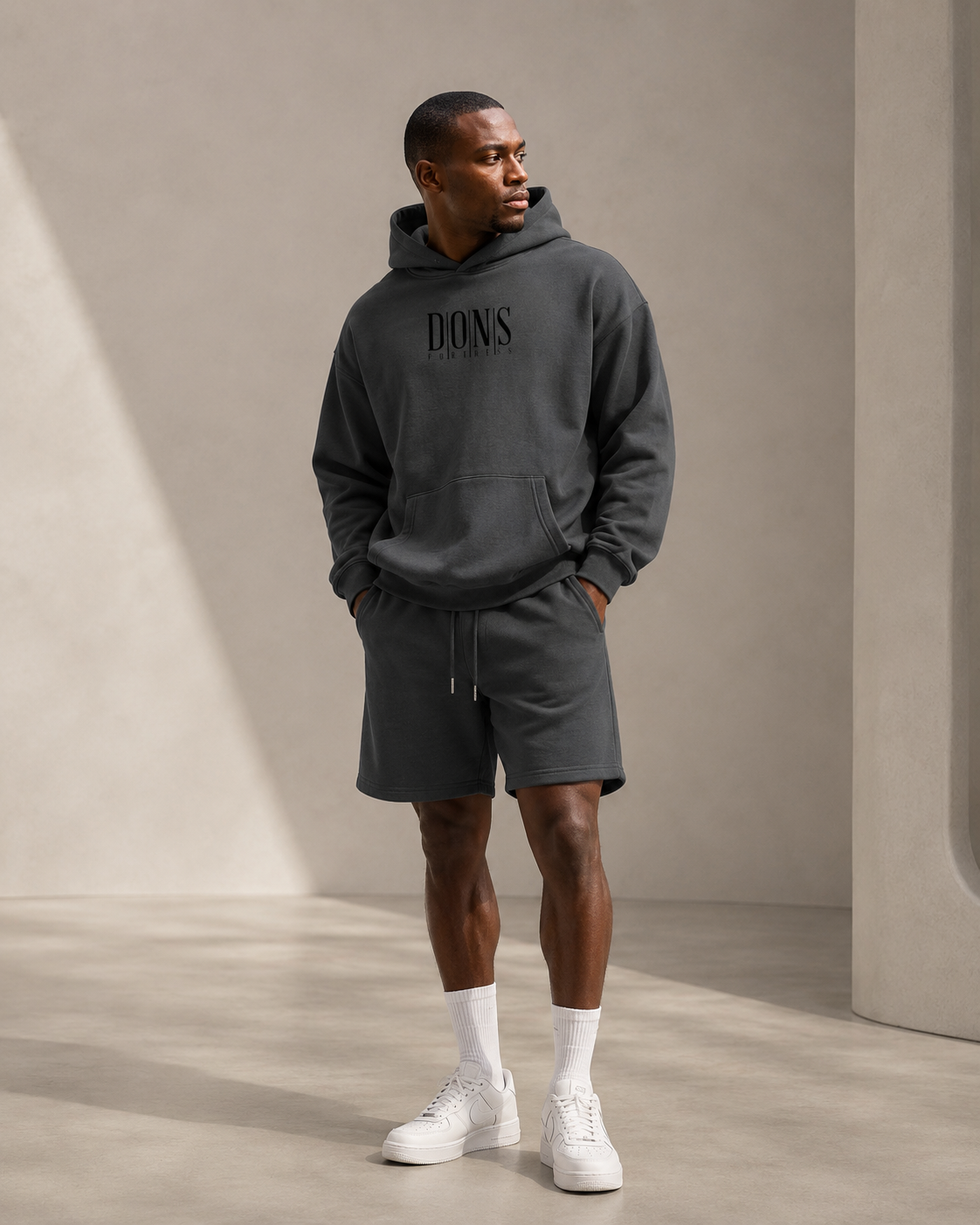 Man wearing a dark gray hoodie and shorts with branding, standing against a neutral background.