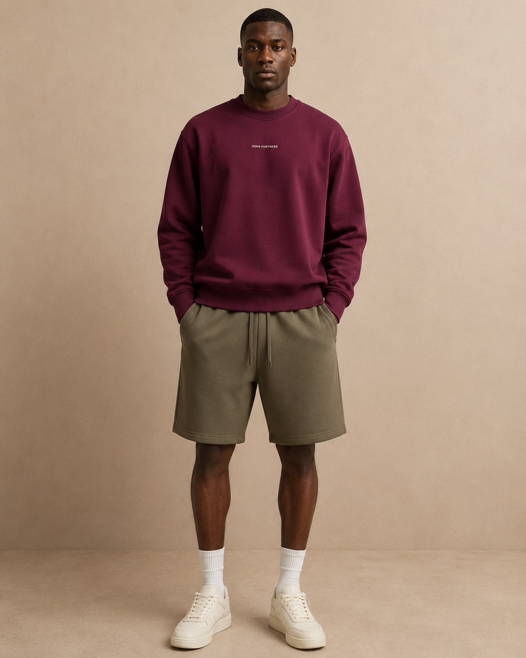 Man wearing a burgundy sweatshirt, beige shorts, and white sneakers against a beige background
