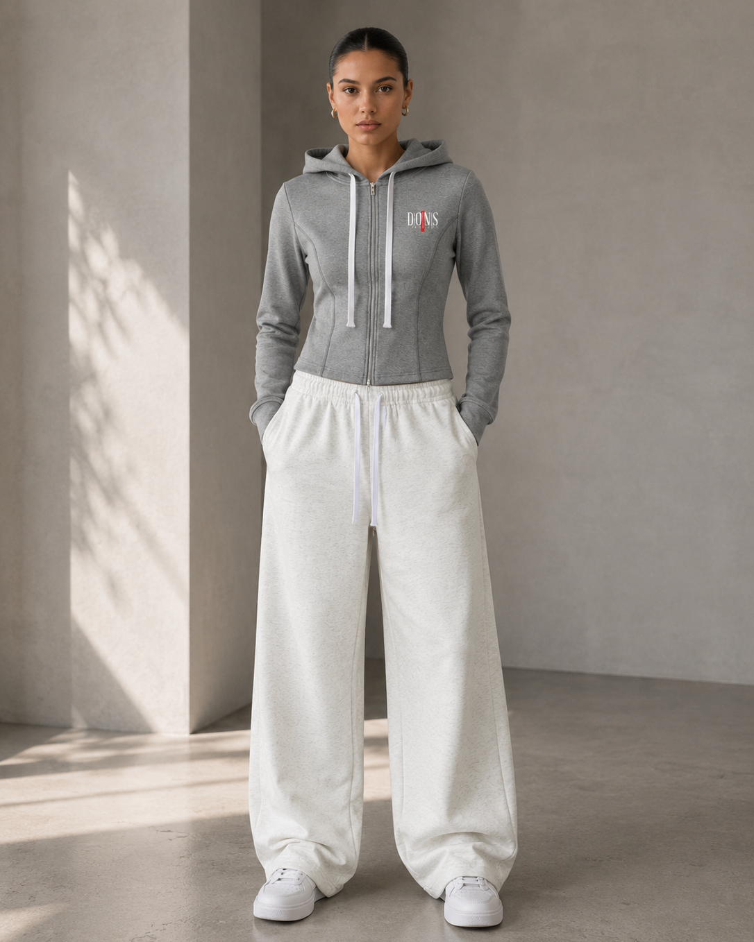 Woman wearing a gray hoodie and white pants standing against a neutral wall.