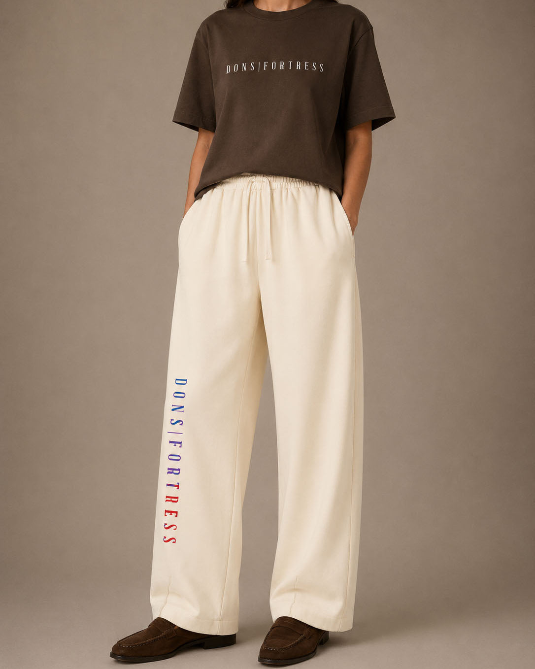 Person wearing a brown t-shirt and white pants with 'DONS FORTRESS' branding on a plain background