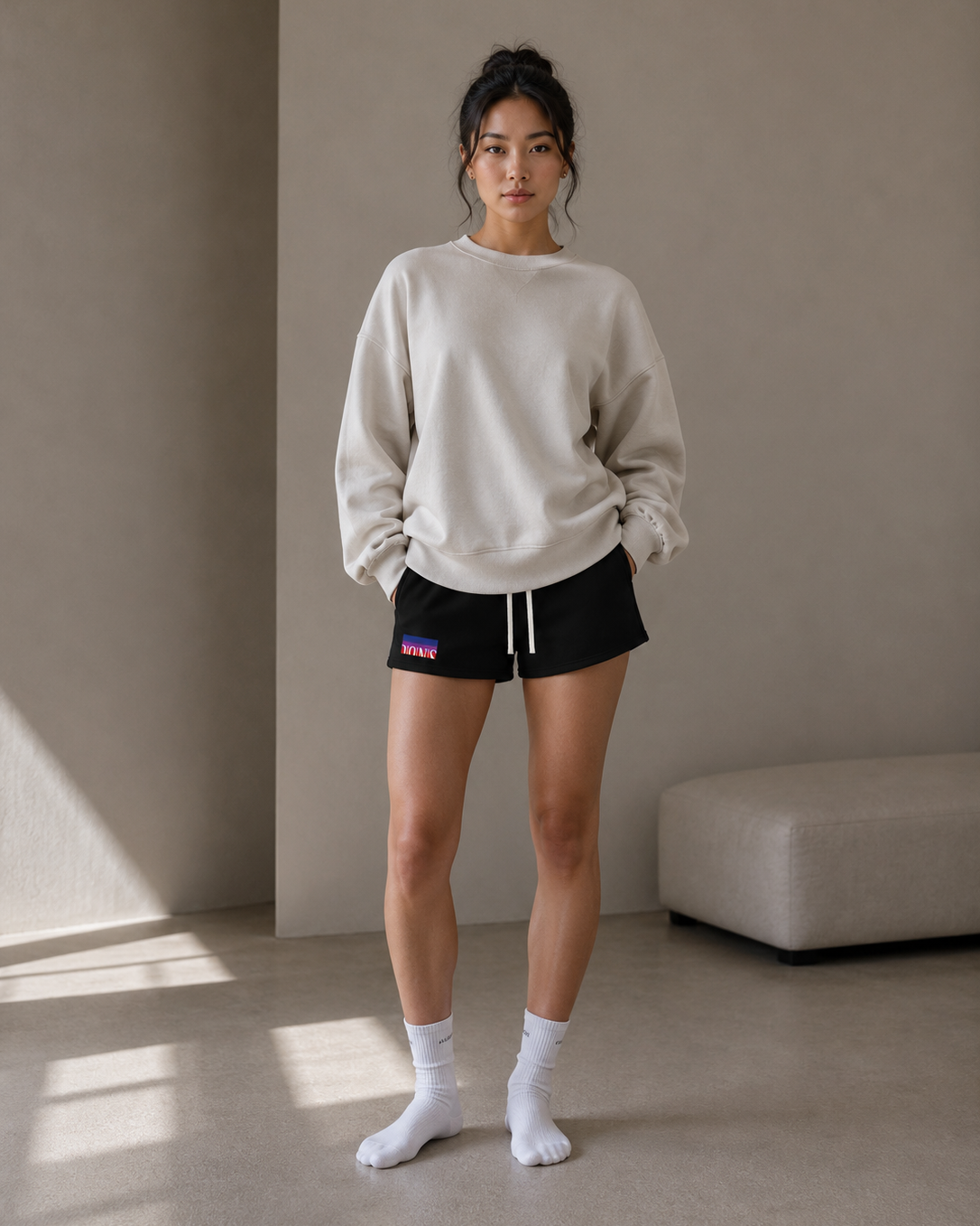 Woman wearing a beige sweatshirt and black shorts with a visible brand logo, standing in a minimalistic room.