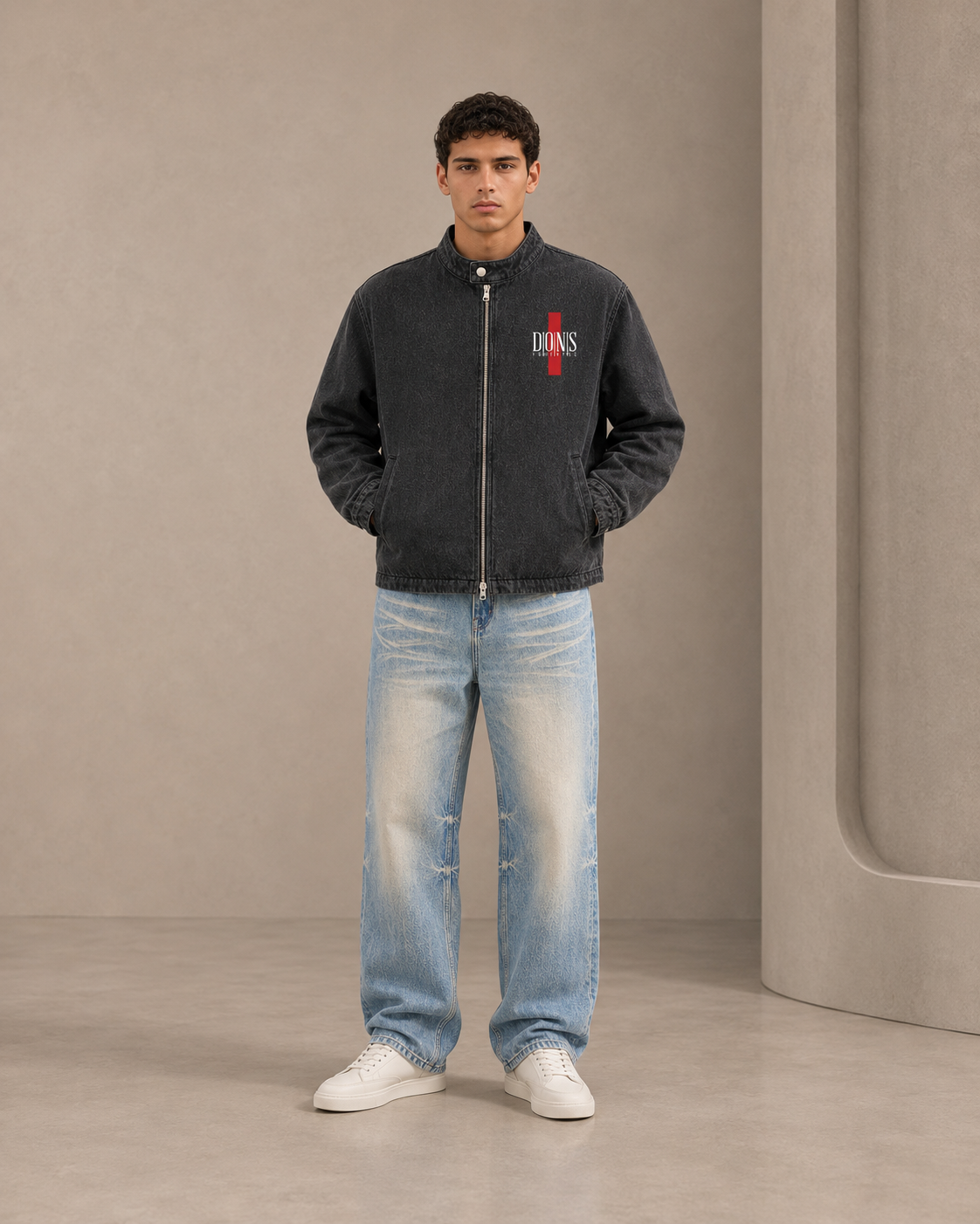 Man wearing a dark jacket with a logo and light blue jeans against a neutral background