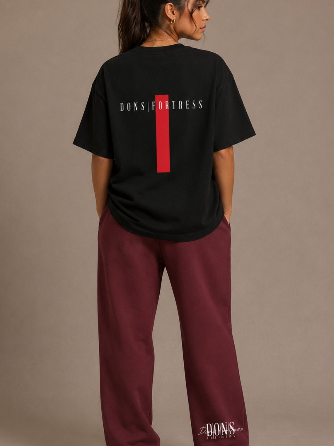 Person wearing a black t-shirt with branding on the back and maroon pants with branding on the side, standing against a beige background.