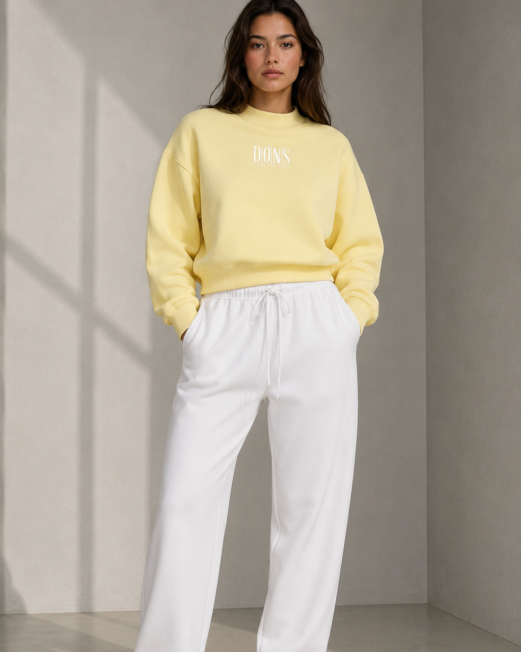 Woman wearing a yellow sweater and white pants standing against a neutral wall.