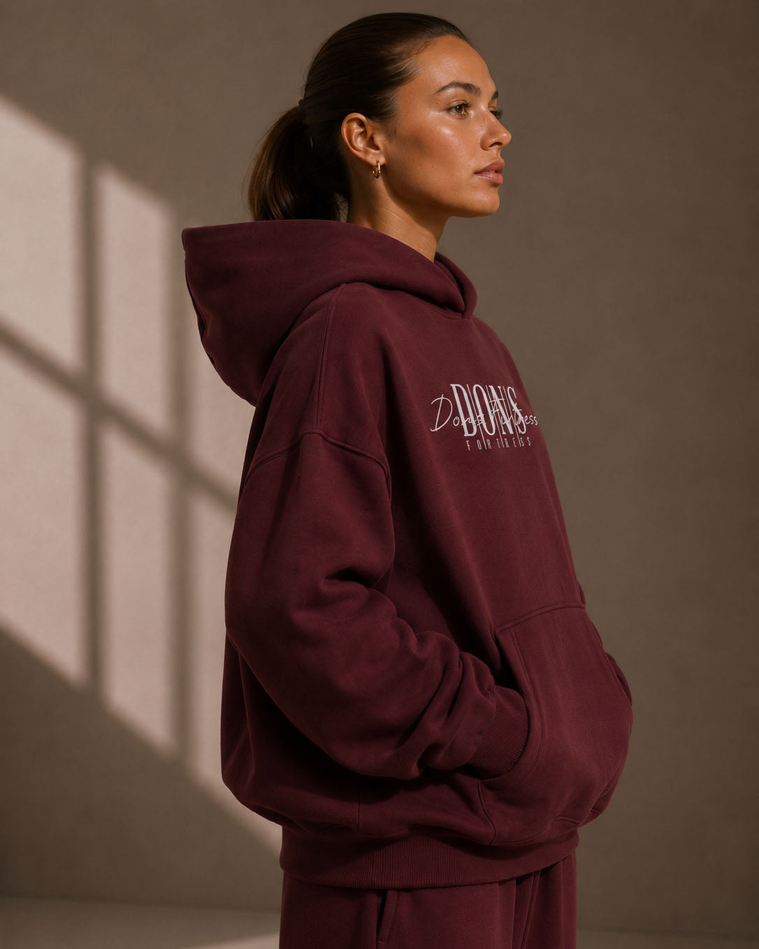 Person wearing a burgundy hoodie with a brand logo against a beige wall.