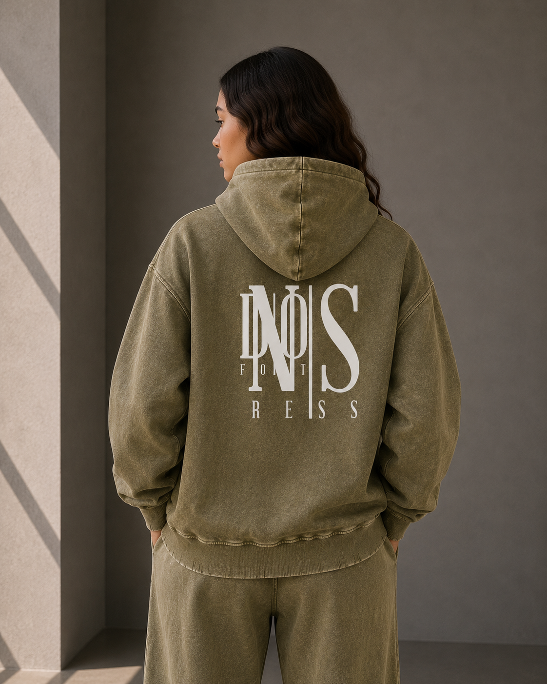 Person wearing an olive green hoodie with a logo on the back against a neutral wall.