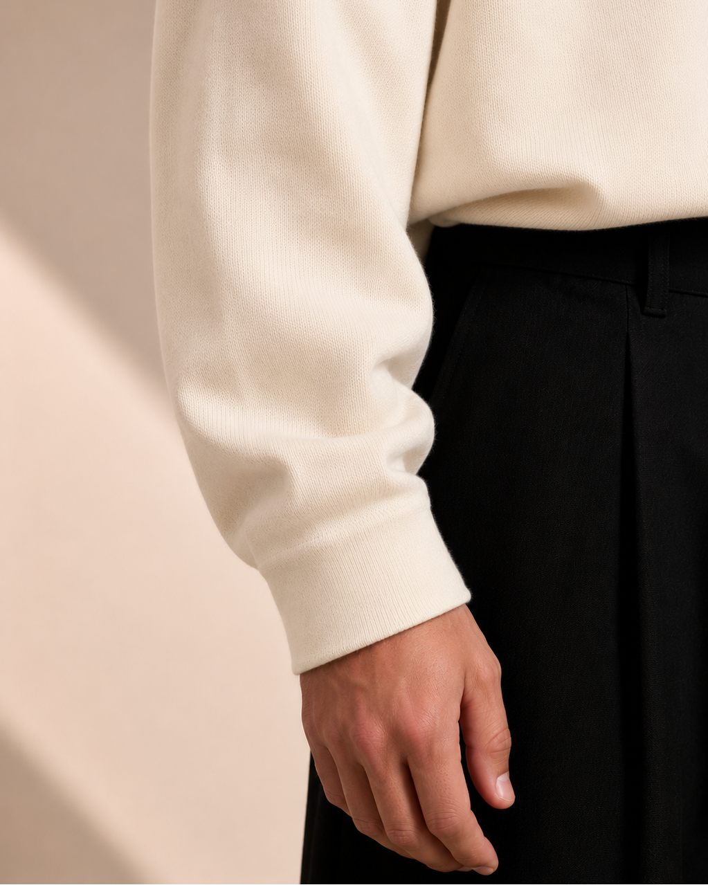 Close-up of a person wearing a beige sweater and black pants against a neutral background