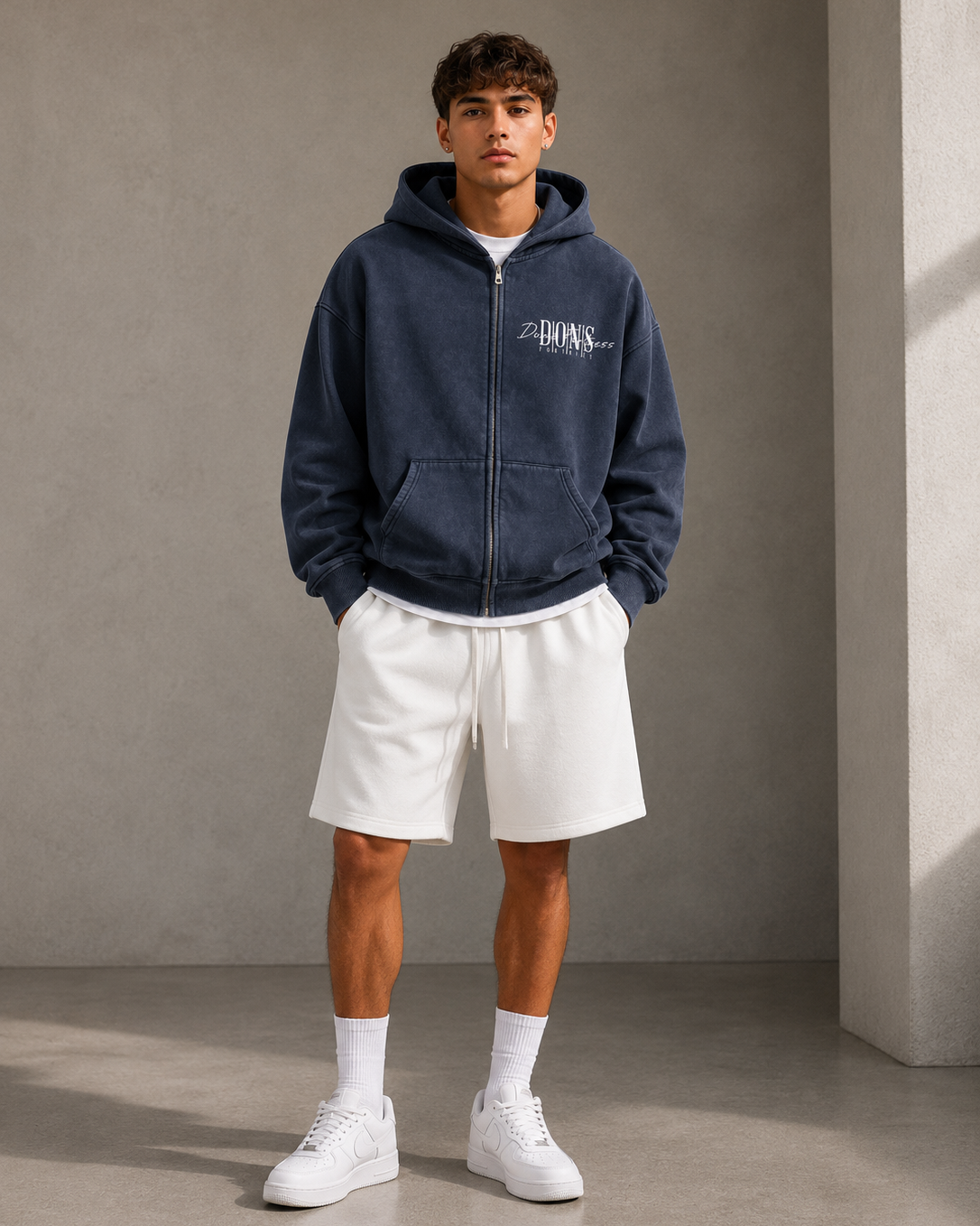 Man wearing a navy hoodie with a brand logo, white shorts, and white sneakers against a gray background