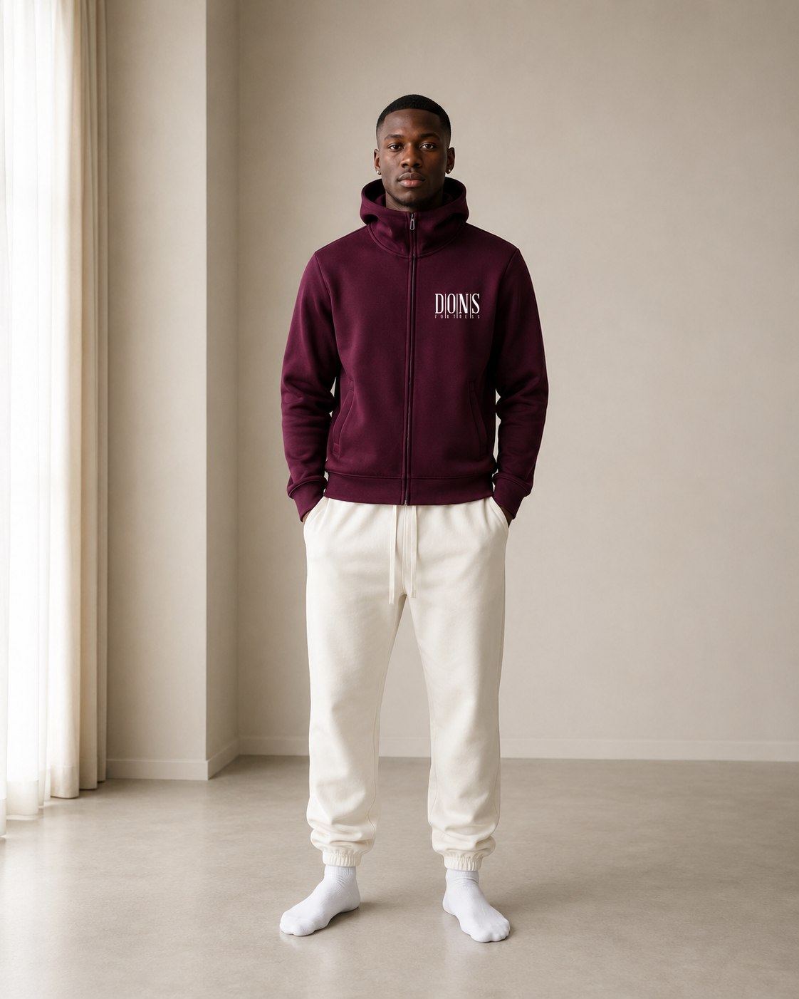 Person wearing a burgundy hoodie with branding and white pants in a minimalistic room.