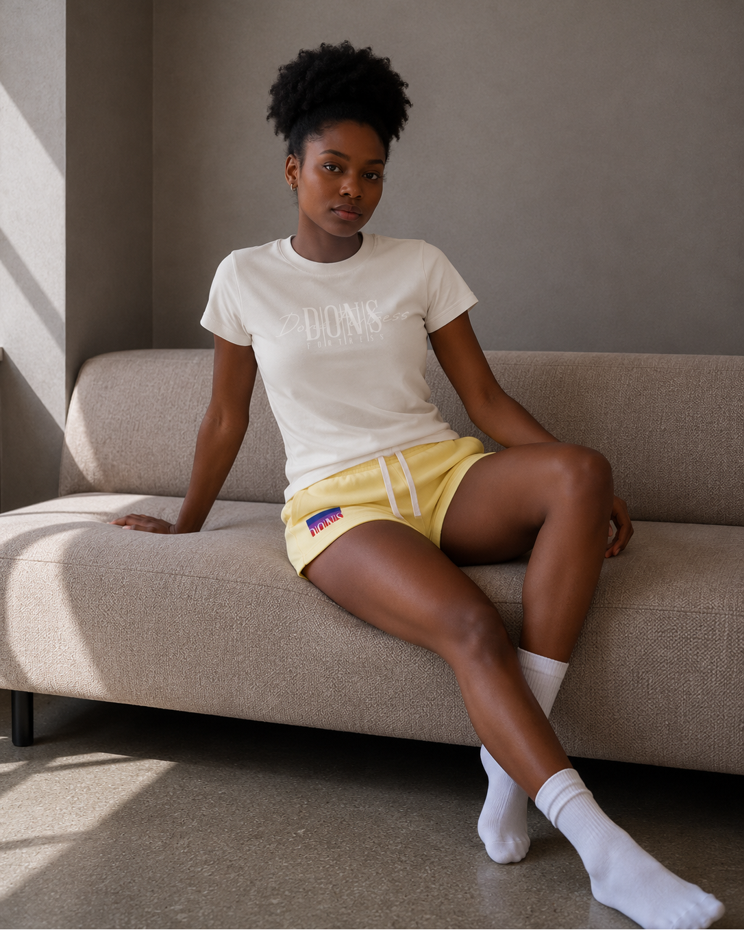 Woman sitting on a couch wearing a white t-shirt and yellow shorts.
